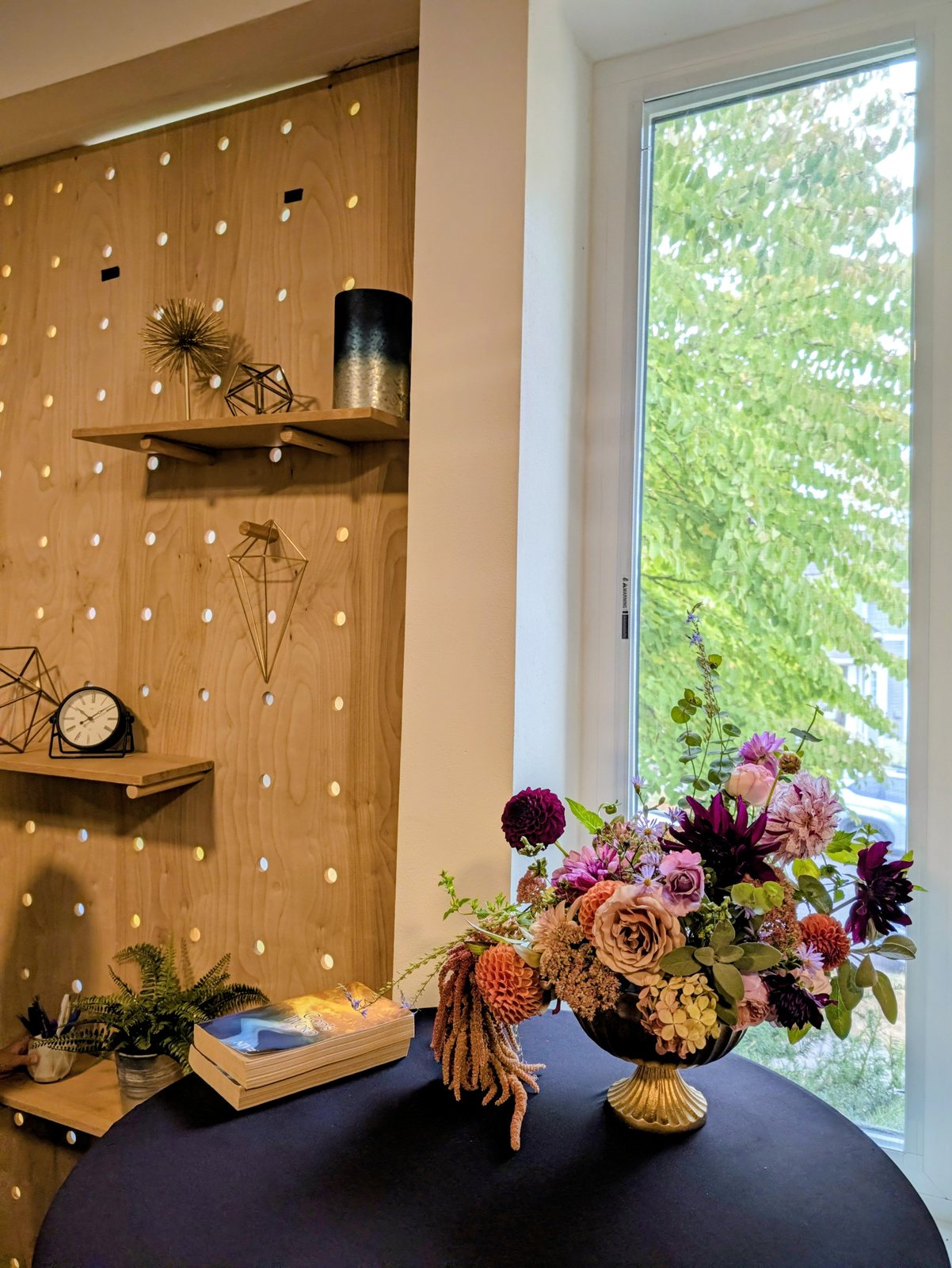 Gold compote centerpiece of dahlias, garden roses, amaranthus, and hydrangea, styled on a navy table beside a sunlit window