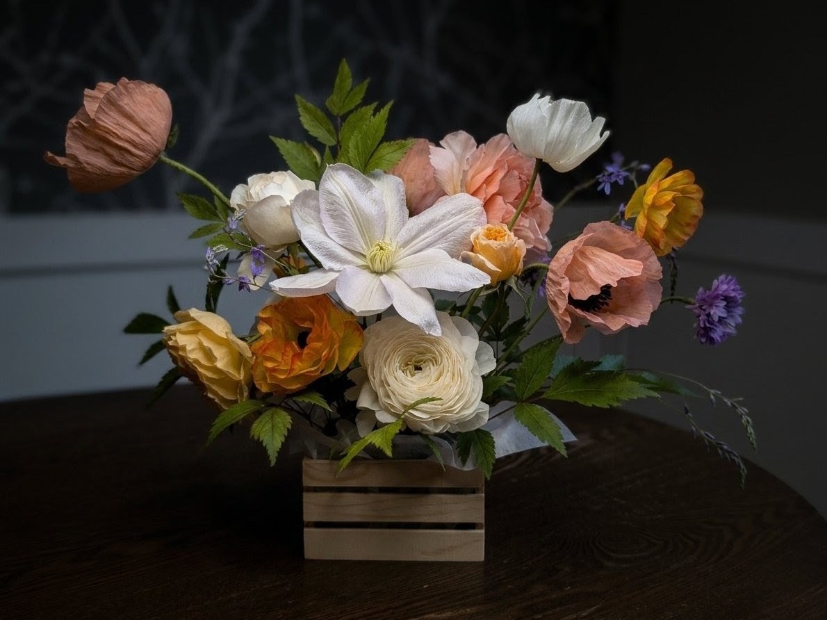 Spring arrangement in a wooden crate with apricot poppies, white clematis, ranunculus, garden roses, and cornflowers